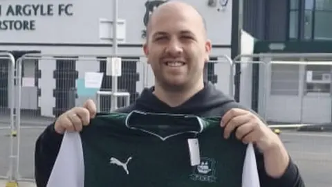 A person holds up a Plymouth Argyle shirt outside Home Park stadium, with the Plymouth Argyle FC Superstore visible in the background. 