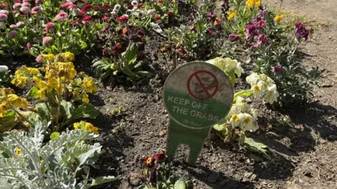 BBC/JO BURN A keep off the grass sign in a flower bed at Westgate Gardens, surrounded by brightly coloured spring blooms.
