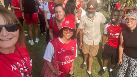 Jen Craft A woman wearing black sunglasses on the left of the photo while she takes a photo. Also in the frame is three woman and two men. One woman is wearing a red top and a red sunhat, while another woman behind her is smiling and wearing a red blouse. Next to her is an eldery man wearing a turban, a buttoned up shirt, long shorts and loafers. Next to him is another man wearing a red shirt and black shorts. There is a woman at the end of the photo on the right side and she is wearing floral trousers and a black top. They are standing outside on the grass and there are a lot of people outside behind them. 