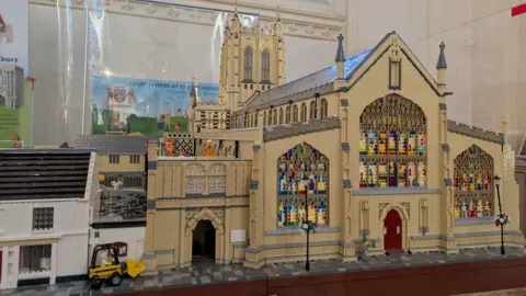 The outside of a model Lego replica of St Edmundsbury Cathedral with a spire and tower and stained glass windows.