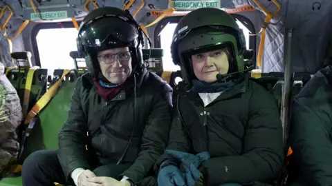Moose Campbell/BBC A woman and man sat in dark clothing with helmets on, on-board a helicopter. Behind the pair are two emegency exit signs.