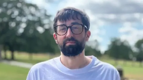 Gemma Dillon/BBC Nick Simpson wearing a t-shirt, has thick black beard, black hair and glasses, stood in park
