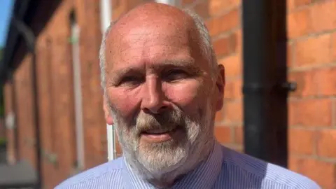 Chairman Bill Anderson has a grey beard and is wearing a blue and white striped shirt. He is smiling beside a large bricked building. 