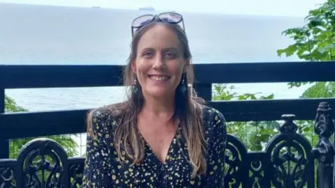 A woman with long hair and glasses on her head. Some leaves and the sea can be seen in the background.