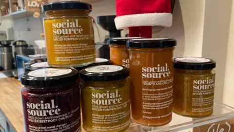 Stacked jars of social source on a wooden counter in Roots cafe. The branding is fairly simple and organic looking, and inside there are various types of chutney. In the background you can see a coffee machine and the bottom of a Christmas elf decoration. 