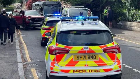 Two police cars and a van block a village road. A maroon pick-up and a small bus can be seen in the background. On the right hand side of the image a police officer is talking to a man wearing a dark-coloured t-shirt and matching shorts. on the opposite side of the road a young boy and girl can be seen walking along the pavement with their backs to the camera. 