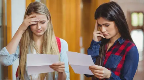 Two girls stand in a school corridor looking anxiously at sheets of paper they are holding. One hard blonde hair and the other had dark hair. 
