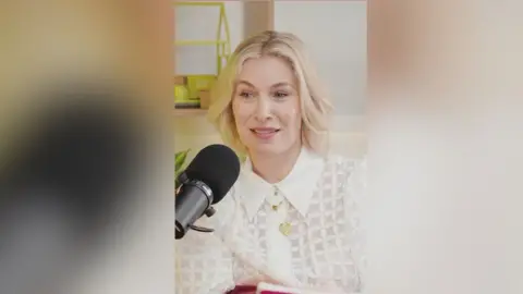 A woman in a white top speaks into a microphone.