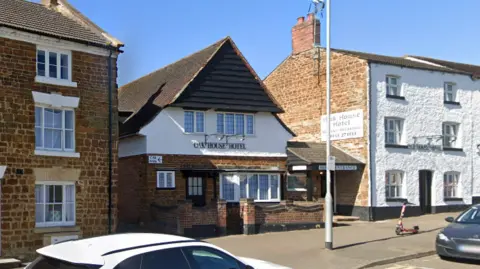 The Oak House Hotel which stretches across three buildings. A white and black car are also parked on the street and can be seen in the picture.