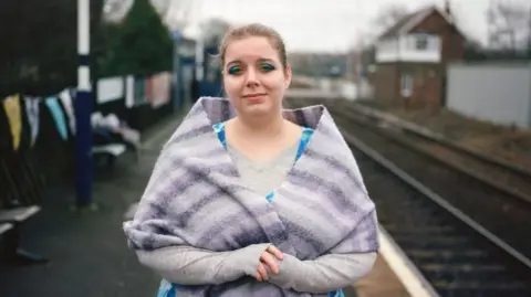 Joanne Coates Head and shoulder shot of Lizzie Lovejoy, who has tied-back brown hair. They are clasping their hands and wearing a striped purple shawl over a long-sleeved mauve top. Behind, and out of focus, can be seen a railway track with an old signal box to one side and a fence with bunting on the other.