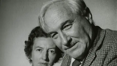 A black and white photo of Louis with his second wife Mary Leakey. He is close to the camera and has a small moustache, light coloured hair and is smiling with his mouth closed. His wife has short dark and curly hair. She further behind Louis and also looking at the camera. 