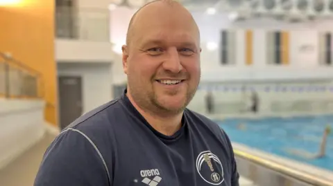 Kate Bradbrook/BBC Andy Sharp wearing a navy Northampton Swimming Club. He is looking directly at the camera with a broad smile and people can be seen swimming in a blurred background