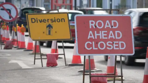 BBC Road closed sign and yellow diversion sign as cars drive past.