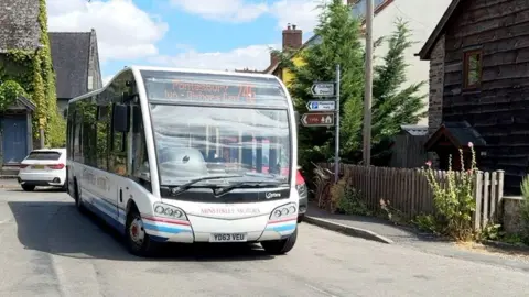 A white bus with red and blue stripes on it drives down a road with houses and trees and a road sign behind it.
