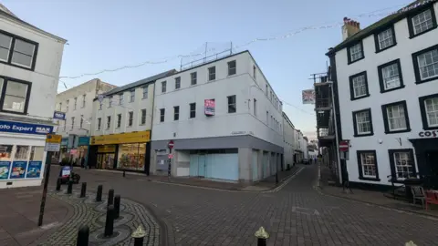 The three-storey former Halifax branch on King Street in Whitehaven town centre. The building, on a corner site, is a white retail unit with boarded up windows. It sits on a crossroads of cobbled streets. A red "to let" sign is displayed on an upstairs window.