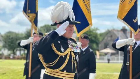 The lieutenant governor saluting. He is in a black and gold tunic and traditional feathered bicorn hat. Standard bearers line up behind.