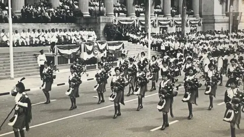 Handout A black and white photo of Singaporean girls performing in a marching band at an event. Officials and the Singapore flag can be seen in the background