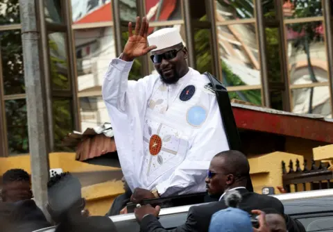 Luc Gnago / Reuters Guinean leader Mamadi Doumbouya in a smart, embroidered white boubou and dark sunglasses waves after submitting his candidacy at the Supreme Court a Conakry, Guinea, Monday 3 November 2025.
