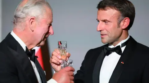 King Charles and Emmanuel Macron holding glasses while dressed in tuxedos