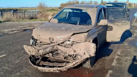 A dark-coloured Honda car caked in mud, its front bumper hanging off, parked by a roadside in the Fens.