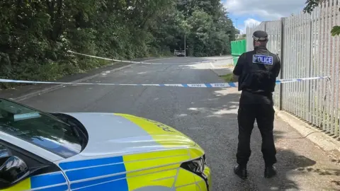 BBC A police officer is standing near a police car with his back to the camera, next to a section of road that has been cordoned off using two long pieces of police tape. There are trees and a pavement on the left side and a metal fence on the other.