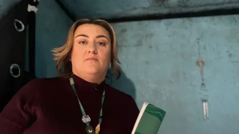 Emma Baugh/BBC News A female housing officer in front of a blue wall with black mould and water running down the walls.