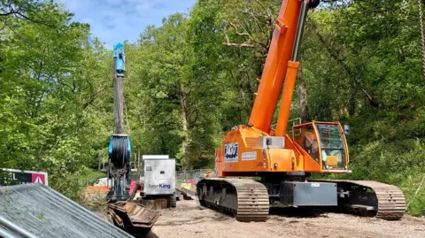 Gloucestershire County Council Two large machines are pictures as roadworks are taking place on a country road. There are trees on either side of the machinery.