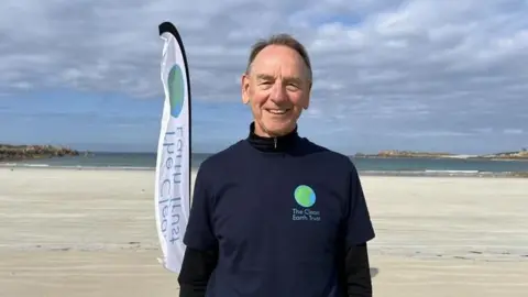 Paul Bugden stands on a sandy beach under a partly cloudy sky, wearing a navy T‑shirt with The Clean Earth Trust logo. A branded flag is nearby, with calm sea, distant rocky headlands, and open shoreline visible in the background.