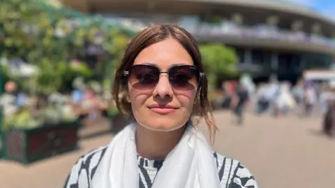 Amara Basit with the tennis club building behind her. She is wearing sunglasses and a white scarf over a black and white top. Her hair is tied back.