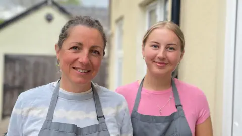 Picture of two women wearing aprons. On the left stands a woman aged 49 smiling at the camera. She has dark brown hair, blue eyes and smiles at the camera. On the right, a woman in her late teens stands smiling at the camera. She has blonde hair and blue eyes and wears a pink t-shirt.