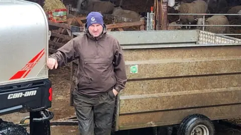 Mark Mather Mark Mather is in his 30s and is standing in the rain wearing a blue hat and a brown jacket. His hand rests on a trailer, behind him 