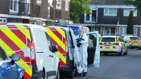 EDDIE MITCHELL A forensic examination officer in a white suit stands beside a row of police vehicles.