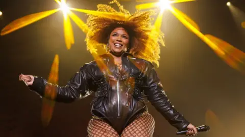 Getty Images Lizzo on stage in a black leather jacket and fishnet tights, in front of bright yellow lights