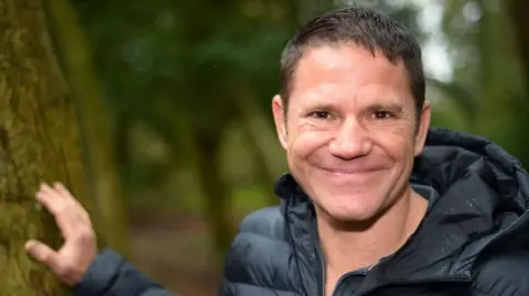 A man with brown hair is smiling at the camera. He is wearing a black puffer jacket and has one hand on a mossy tree beside him