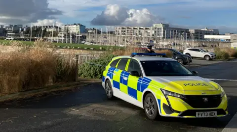 A police vehicle and cordon in place near a marine. Boat masts can be seen in the background as well as tall buildings. The sky is cloudy.