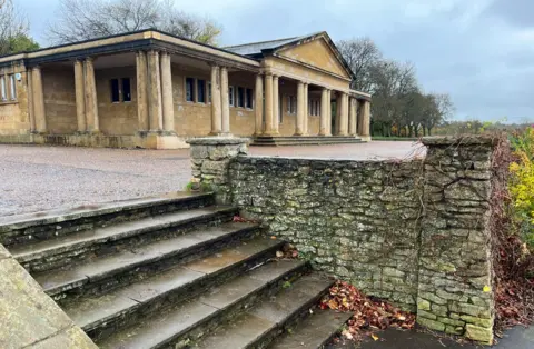 Shaun Whitmore/BBC A sandstone pavilion with many columns supporting the covered area in front of the one-storey building. It has a pointed gable roof a the front, where steps lead up to the terrace. Steps lead down from a paved area at the front, which have a stone wall.