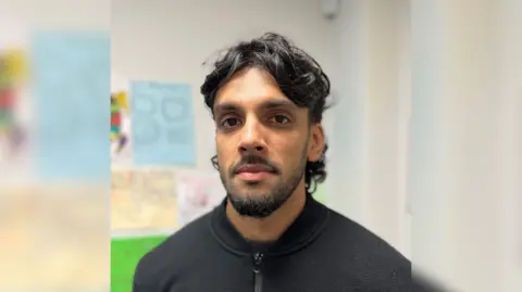 A man with short black hair and a short beard looks towards the camera. The background is out of focus but some paper with schoolwork can just about be made out hanging on the wall behind him.