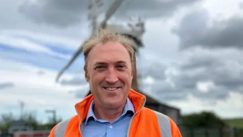Owen Sennitt/BBC A man wearing an orange high vis jacket and shirt smiles towards the camera, with a blurred windpump visible in the background