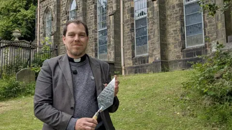 A man with dark hair in a grey jacket and jumper with a vicar's collar, standing on a grassy hill with a church building behind him, holding a shiny, engraved trowel