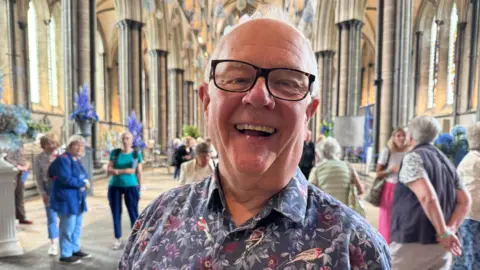 Michael Bowyer wearing a shirt with flowers and birds on it smiling at the camera. He is standing in the main cathedral hall.
