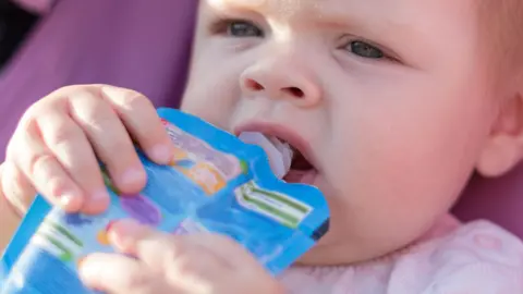 A young toddler holds a blue-coloured food pouch to its lips and is eating from it while sitting in a pram