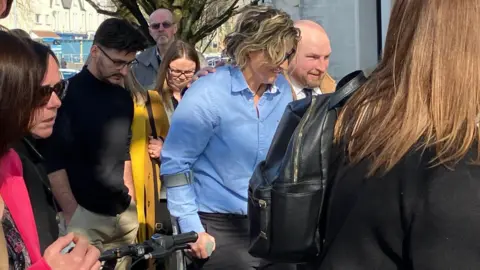 Nerys Lloyd, who has short, blonde, wavy hair and is wearing a blue shirt and sunglasses, holding onto a crutch and walking. She is surrounded by dozens of people and is smiling and looking down at the ground.