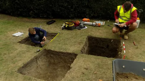 East Herts Council An archaeologist squatting in a rectangular shaped pit making measurements. Another, wearing a high -vis vest, making notes on a clipboard next to two further rectangular shaped pits.