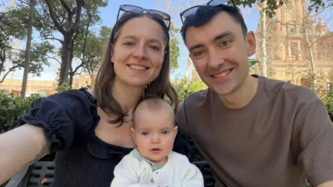 A woman with a black shirt is sitting next to a man in a brown jumper. There is an infant sitting in between the couple.
