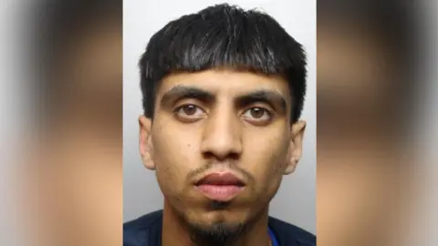 A mugshot of a man who has black hair and brown eyes. He looks straight at the camera, unsmiling.