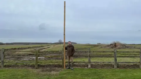 A man wearing wellingtons and a brown coat is standing in front of open countryside and holding a 14.7ft (4.5m) high stick in his right hand.