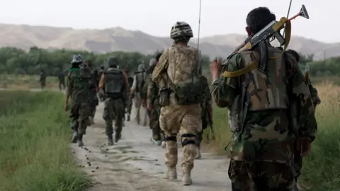Getty Images British Army soldiers patrol with Afghan National Army soldiers with their backs to the camera, walking down a path through a grass field with mountains in the background, in Helmand Province in 2007.