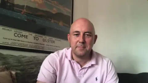 Jonathan Corry is a bald man wearing a pink polo shirt. He is sitting on a sofa. In the background is a plain white wall on the right, and on the left is a poster featuring a picture of boats on a marina. The poster says "Come to Ulster".