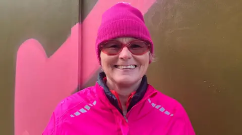Chloe Aslett/BBC A woman with a neon pink jacket, neon pink hat and pink tinted glasses smiled at the camera. Behind her is a wall with a pink arrow painted on it.