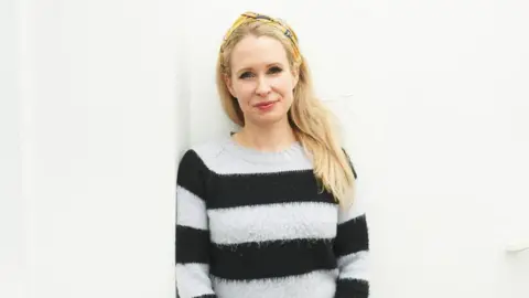 Jonathan Birch/BBC Lucy Beaumont - a woman with long blonde hair and a yellow headband - is leaning against a wall and smiling. She is wearing a white and black striped jumper.